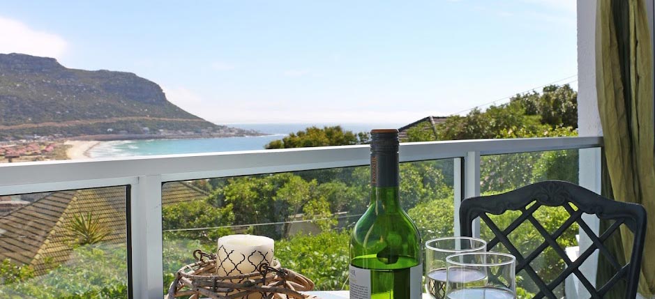 Balcony view from Fish Hoek Bay Apartments with a bottle of wine and two glasses, showcasing the panoramic beach and mountain vistas in Cape Town.
